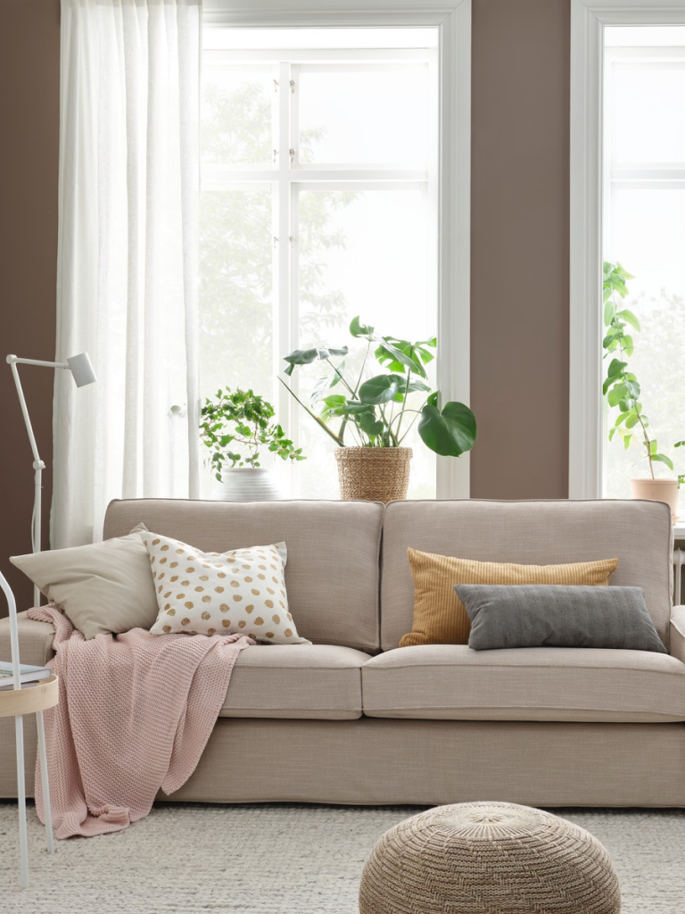 Wohnzimmer mit einem hellen 2-Sitzer-Sofa mit verschiedenen Zierkissen und einer rosa Tagesdecke sowie einer weissen Stehlampe.