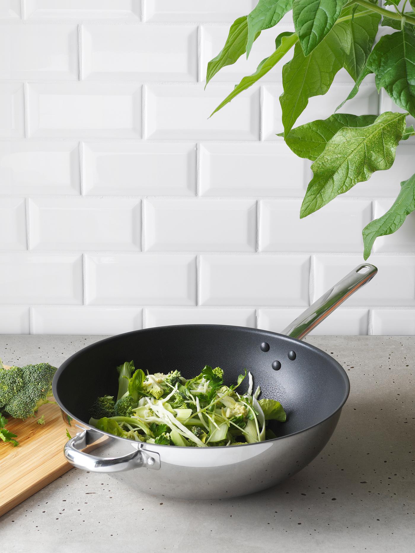 Wok com legumes verdes frescos, como brócolos, em cima de uma bancada de cozinha em cinzento.