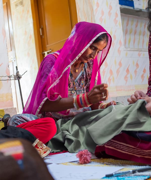 Woman dressed in a colorful dress and sheer shawl sitting on a rug with other women, doing needlework on a piece of fabric.