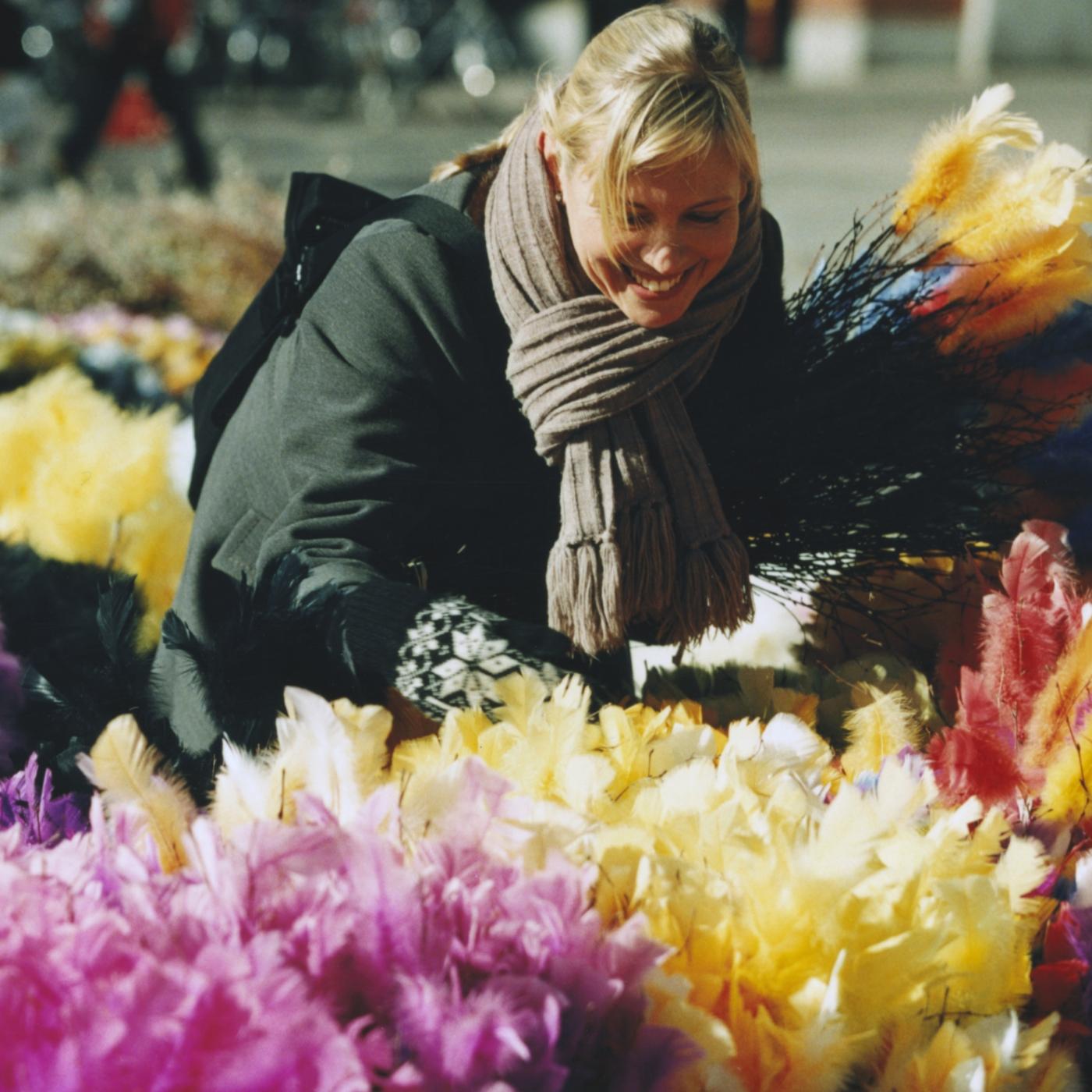 Woman in a green jacket shopping for Easter decoration, surrounded by colourful pink, yellow and red Eastern twigs.