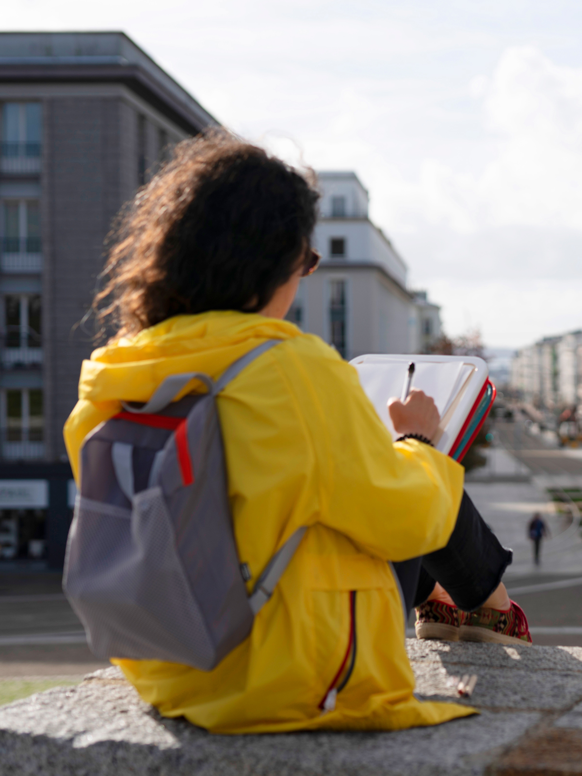 Woman in a yellow raincoat using a MÅLA portable case to draw a painting while out in the city.