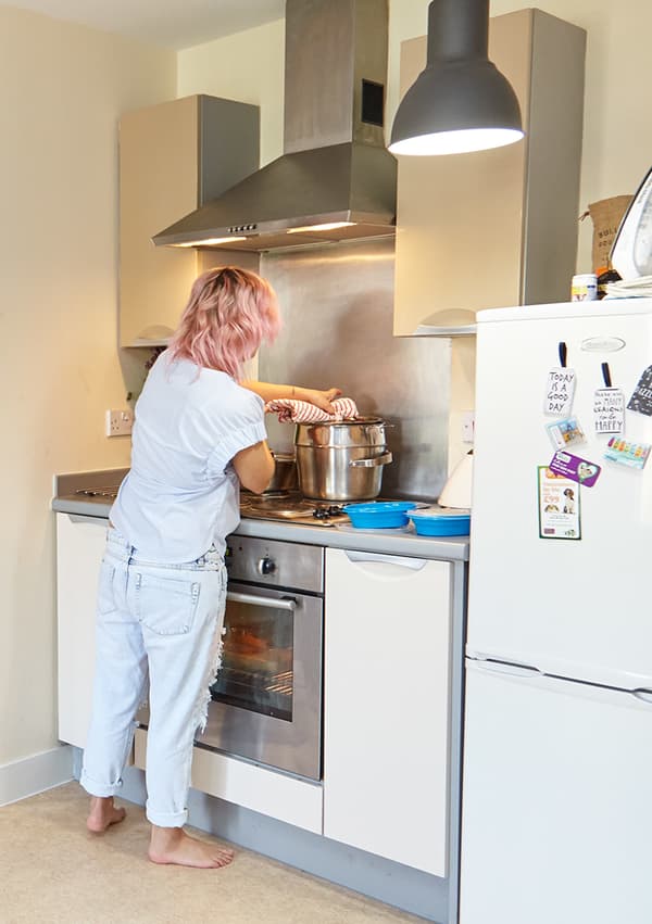 Woman in white clothes and pink hair standing by the stove in her kitchen. 