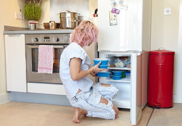 Woman in white clothes kneeling by her freezer, placing plastic containers inside.