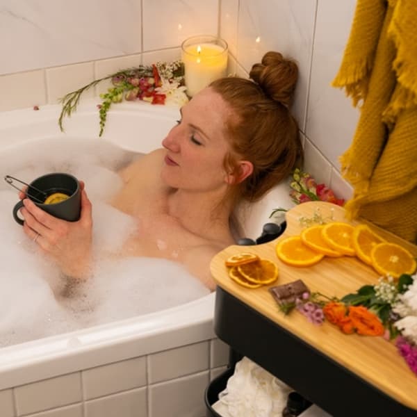 Woman lying in a bubble bath surrounded by candles holding a warm cup of tea with sliced oranges placed on a trolley next to the tub.
