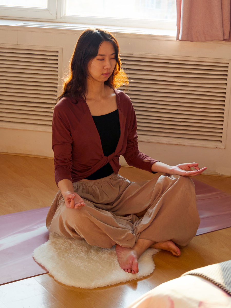 Woman meditating on the floor in her room next to a window, with soft white TOFTLUND rug used for support.