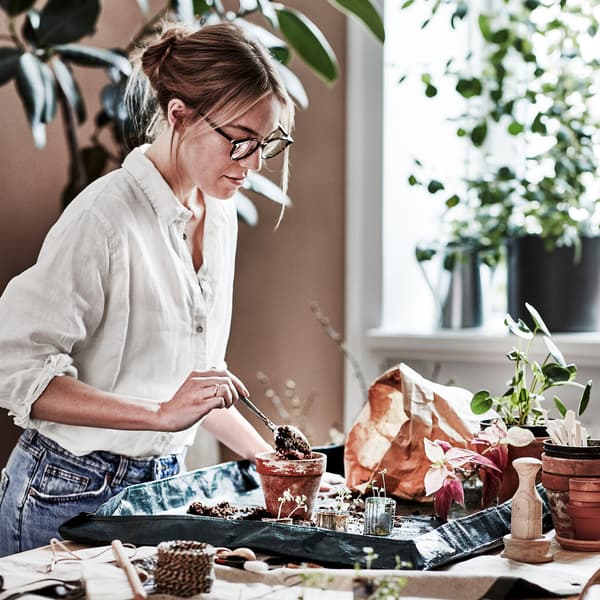 Woman re-potting on a large table, creating a lovely mess, using the plastic sheet of BOTANISK fabric tool storage.
