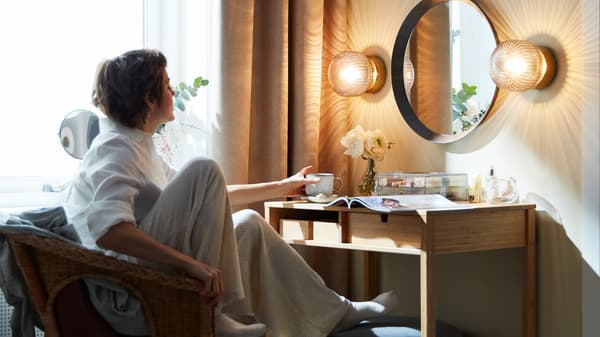 Woman relaxing in an armchair grabbing a mug off a dressing table with a magazine, flower vase and clear box resting on it.