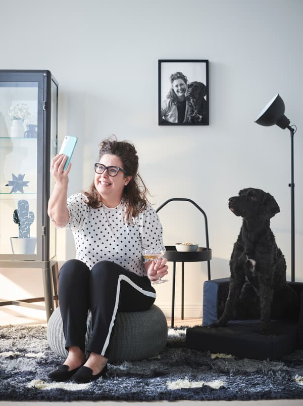 Woman sitting in a living room on a SANDARED pouffe, snapping a selfie of herself and her black dog sitting next to her.