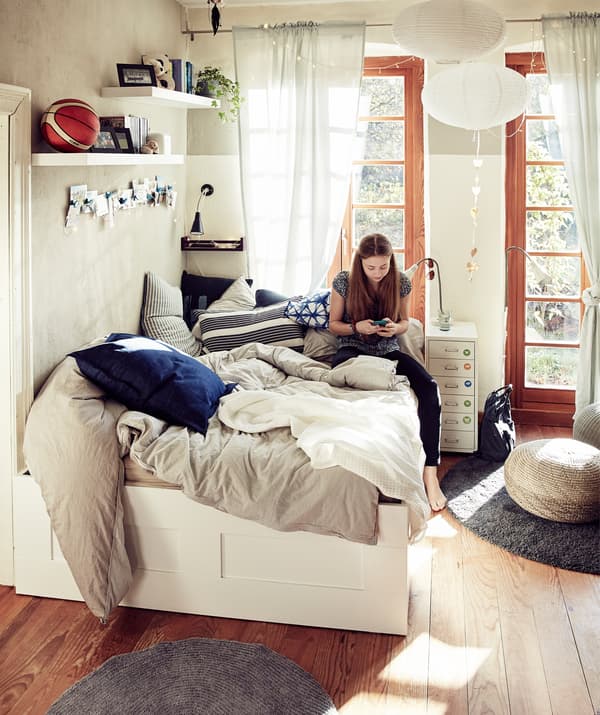 Woman sitting on a double bed with throws and cushions in a bedroom with large windows.