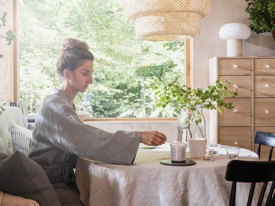 Woman wearing a grey robe sitting at kitchen table with candles beside a brght window with greenery.