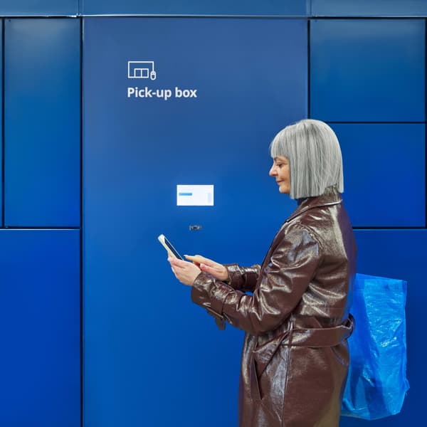 Woman with grey hair in a bown coat standing in front of a wall of dark blue lockers used for pick-up