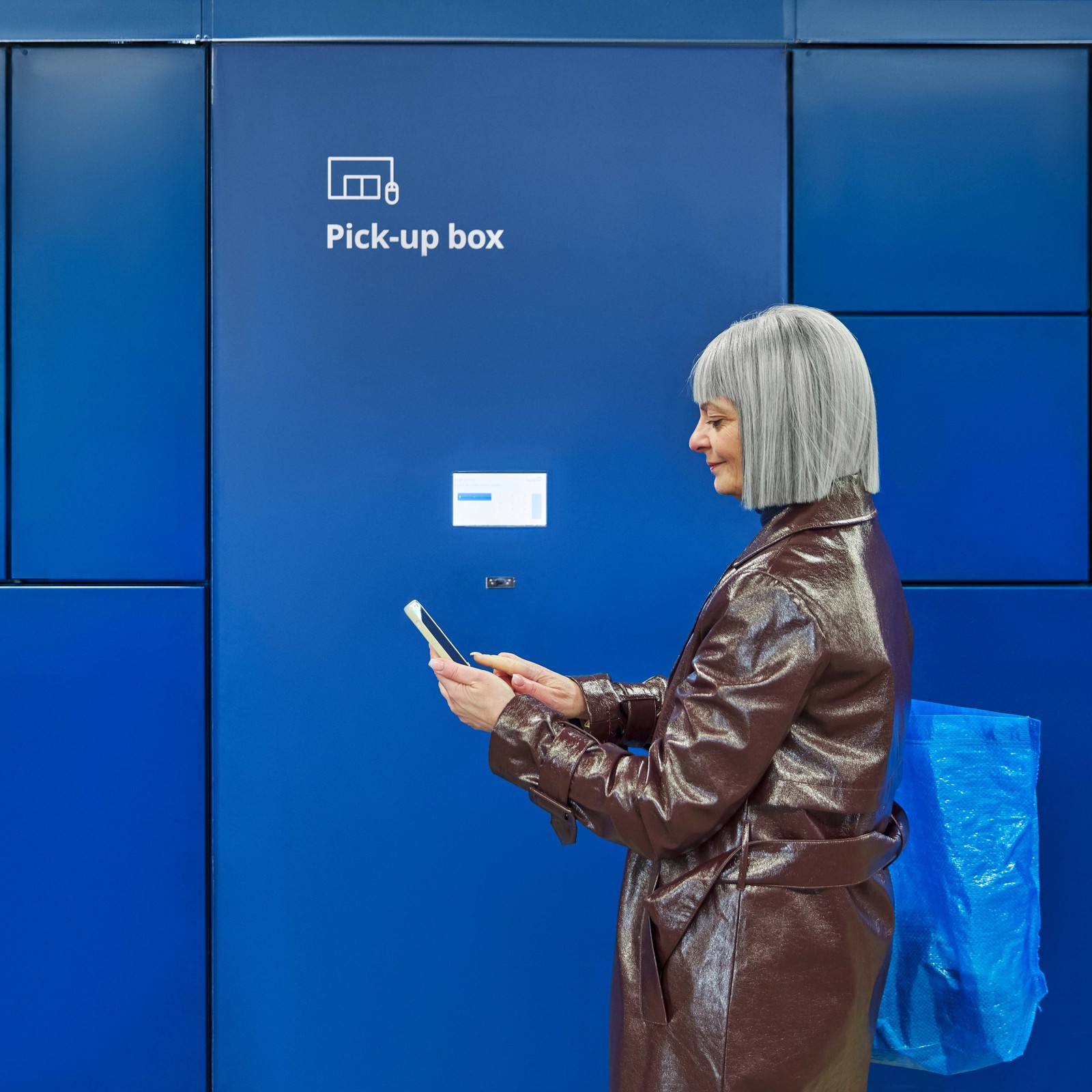 Woman with grey hair in a bown coat standing in front of a wall of dark blue lockers used for pick-up