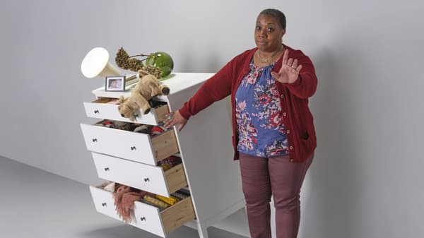 Woman with her hand up expressing to wait while she holds a dresser from tipping over with various items on top and drawers slightly open.