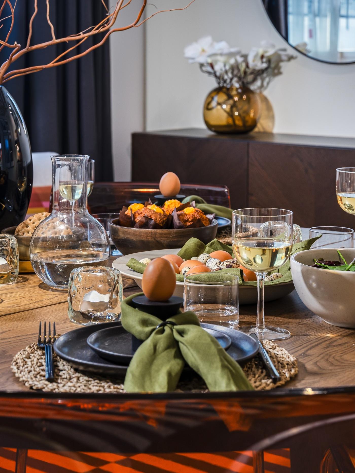 Wooden dining table styled for an Easter gathering with natural tableware, green linen napkins and spring branches in a vase.