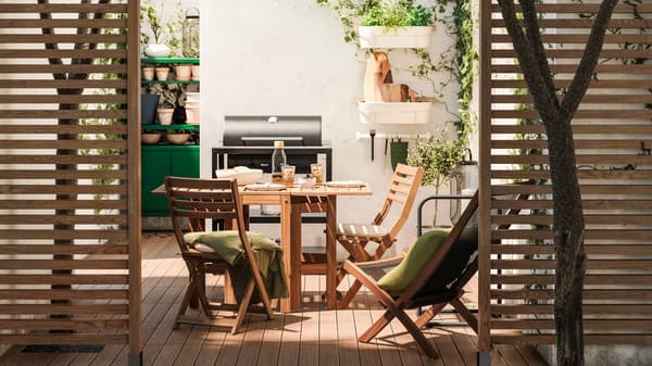 Wooden dividers offer a peek into a patio with wooden furniture, wooden decking, black grill and green storage unit.