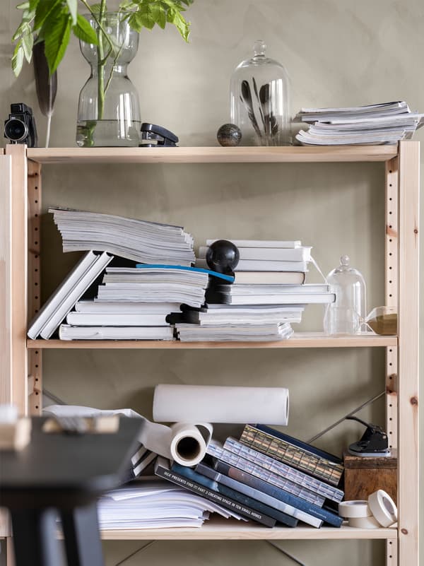 Wooden shelves against a wall with messy piles of books and magazines and glass vases with plants and feathers inside.