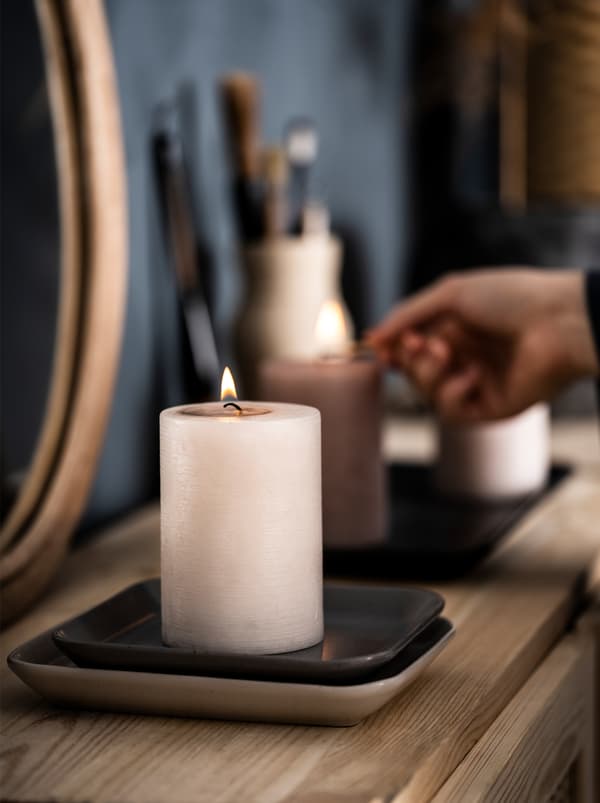 Wooden sideboard in a bedroom with a mirror and decorations. A hand lights two LUGGA scented candles, also placed on it.