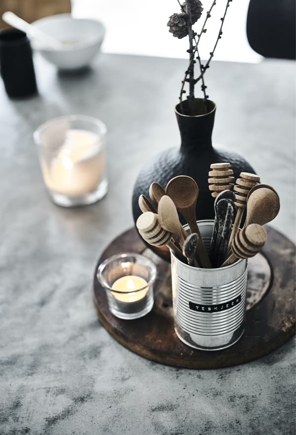 Wooden utensils in a metal can shown together with a black vase and a clear glass tealight holder with a lit tealight.