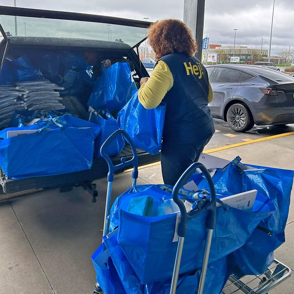 Worker loads large IKEA blue shopping bags into a vehicle outside the store, with carts full of packed bags nearby.