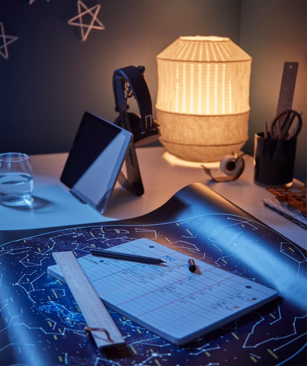 Workspace-style desk with star map and calendar, lit acorn-shaped table lamp and filled tablet stand.