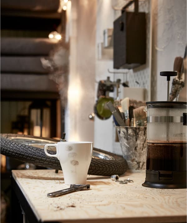 Worktop in a garage-like room, a bike wheel and wrench sharing the surface with a coffee maker and a lightly stained mug. 