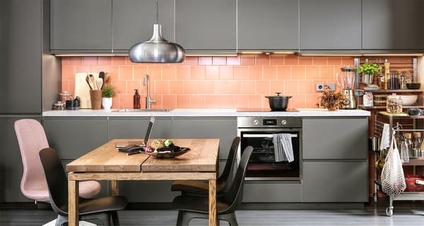 Wyłożona różowymi płytkami ceramicznymi kuchnia z ciemnoszarymi frontami, antracytowymi krzesłami, stołem jadalnianym wykończonym dębową okleiną i lampą wiszącą VÄXJÖ.