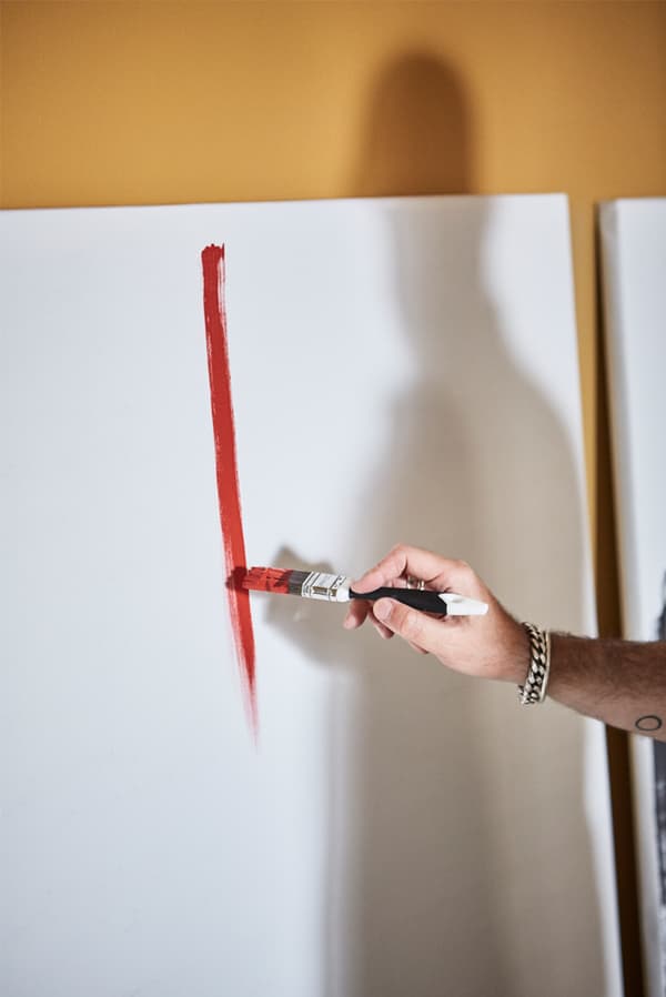 يد رجل تمسك بفرشاة رسم وترسم خطًا أحمر على قماش أبيض مسنود على جدار أصفر اللون بينما يظهر ظل شخص على القماش.