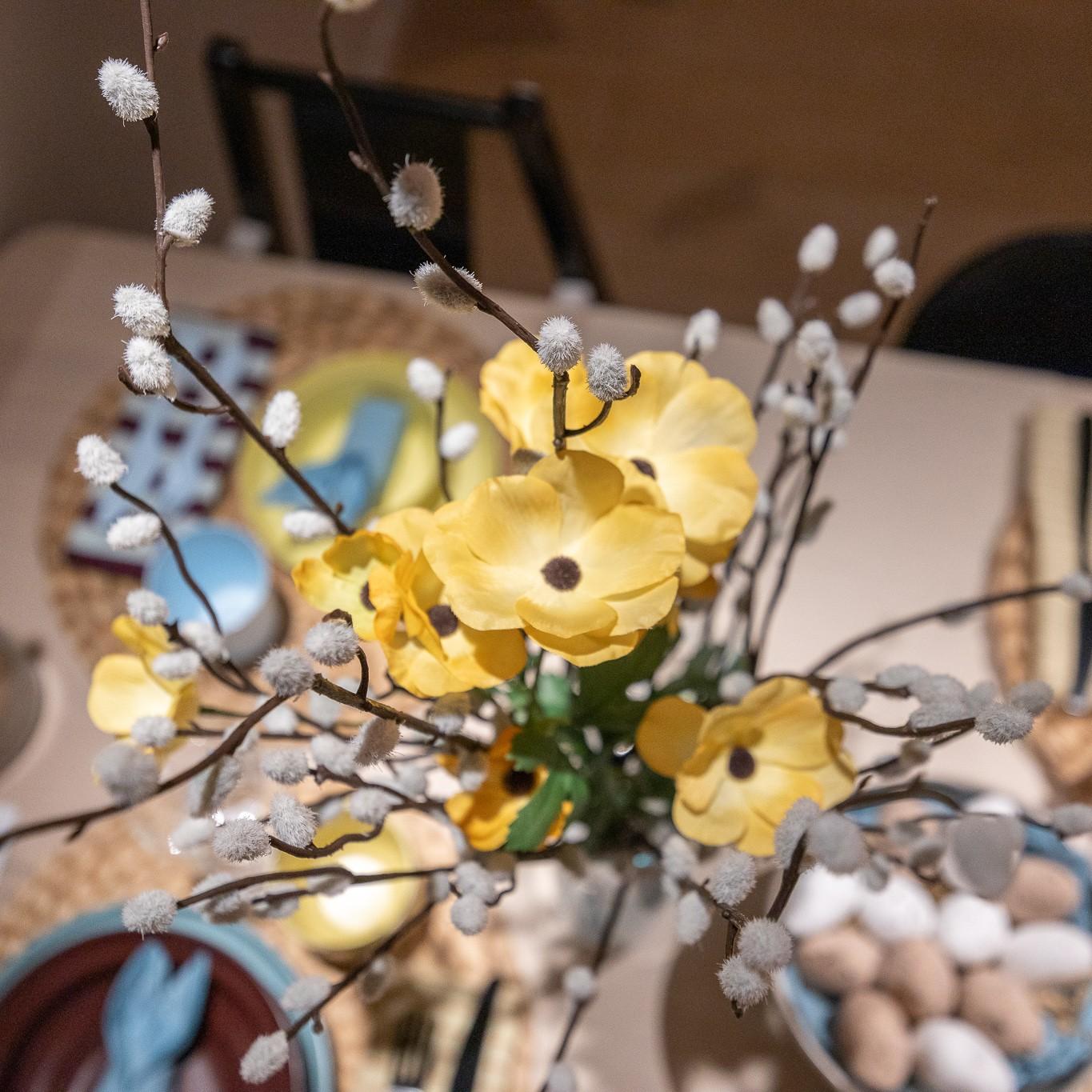 Yellow spring flowers arranged with willow branches, with a blurred view of a festive table setting and decorative Easter eggs in the background.