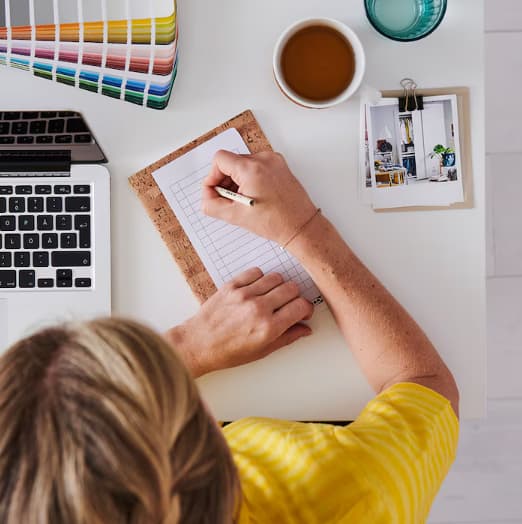 Ylhäältä otettu kuva, jossa pöydällä värinäyteviuhka, näppäimistö ja kahvikuppi. Henkilö tekee muistiinpanoja muistioon.