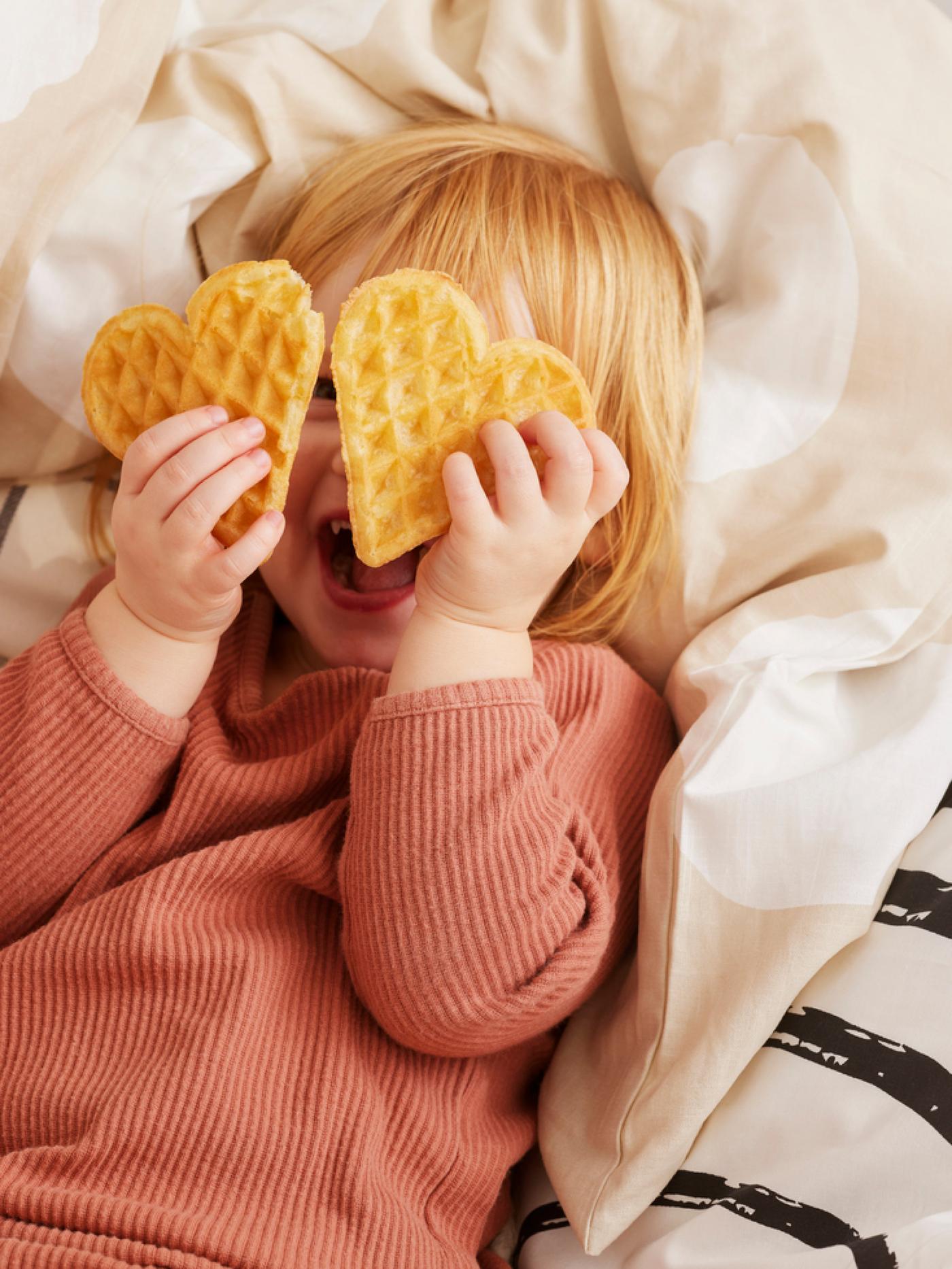 Young child on a bed with two heartshaped waffles in front of her smiling face.