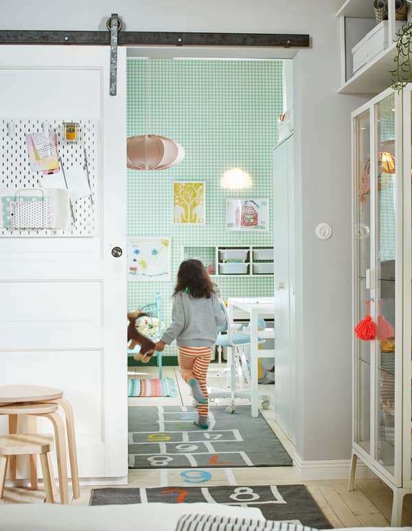 Young child running through the open, sliding door of a white-and-green room furnished with a bed, storage, a desk, and more.