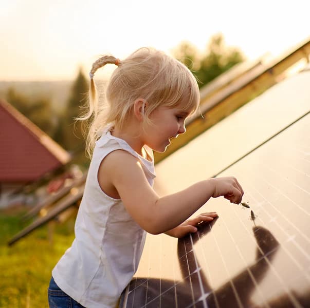 Young girl with blonde hair in a ponytail, wearing a white tank top, touching a solar panel outdoors on a sunny day.