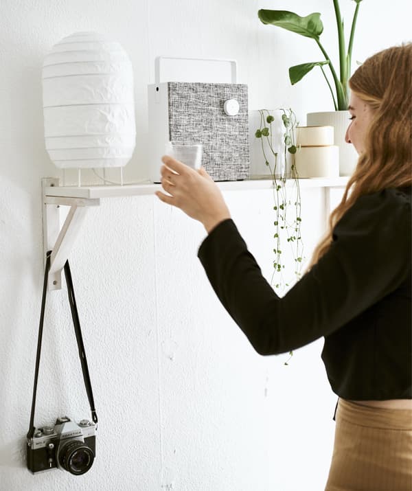 Yvet pose un verre sur une tablette sur laquelle se trouvent une lampe, une enceinte et des plantes.