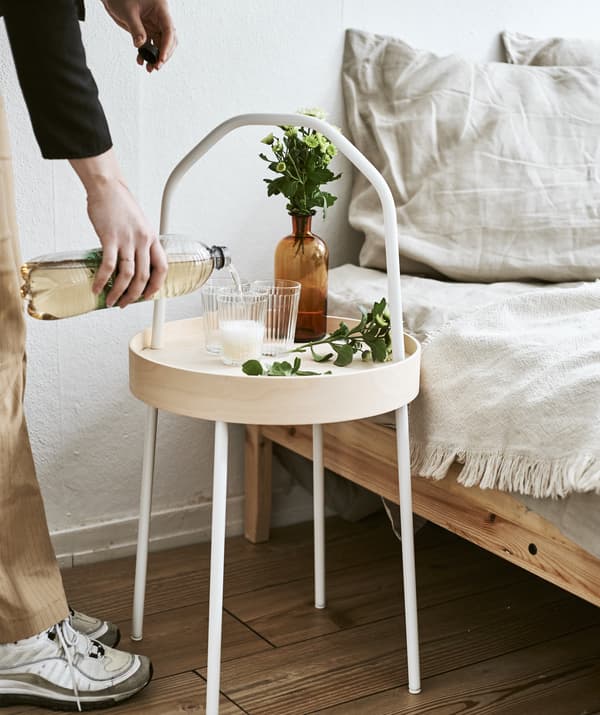 Yvet verse une boisson dans un verre posé sur une table d’appoint en bois clair avec poignée, posée à côté d’un lit.