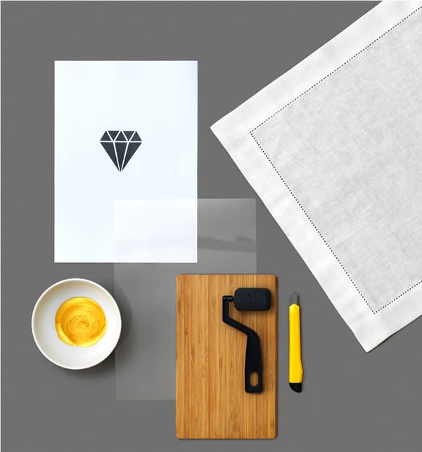 Za izradu šablona za tkaninu u obliku dijamanta i bojanje GULLMAJ ubrusa iz robne kuće IKEA, trebat će ti: drvena daska za rezanje, skalpel, zlatna boja u zdjeli, komad papira s uzorkom koji ćeš izraditi, bijeli ubrus, valjak i list plastike.