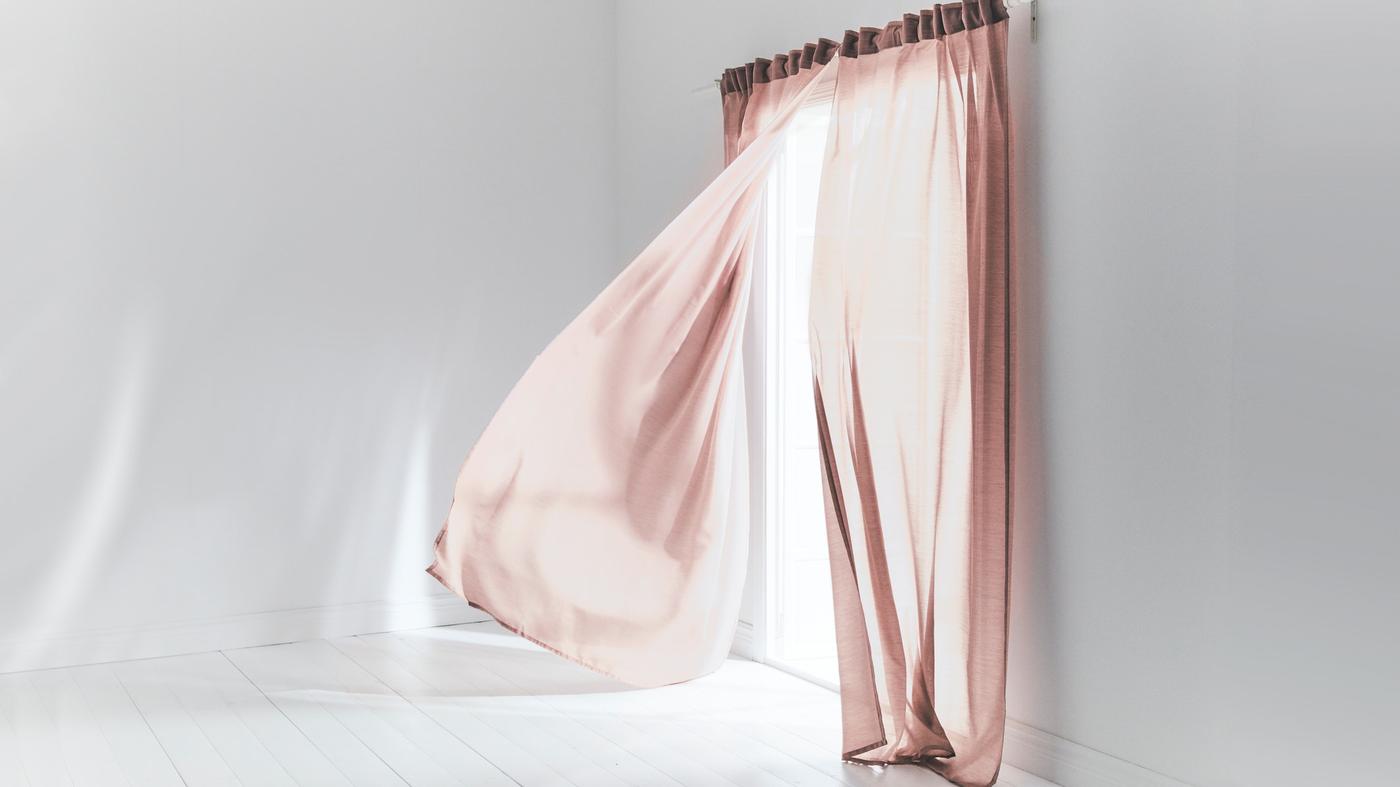 Zachte roze gordijnen waaien bij open raam in lichte kamer met witte muren en vloer.
