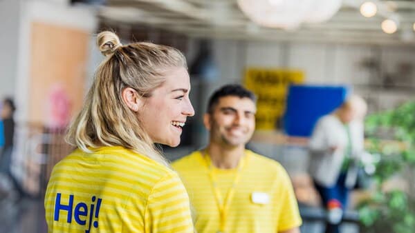 Zaměstnanci IKEA ve žluté uniformě s nápisem HEJ na zádech se usmívají.