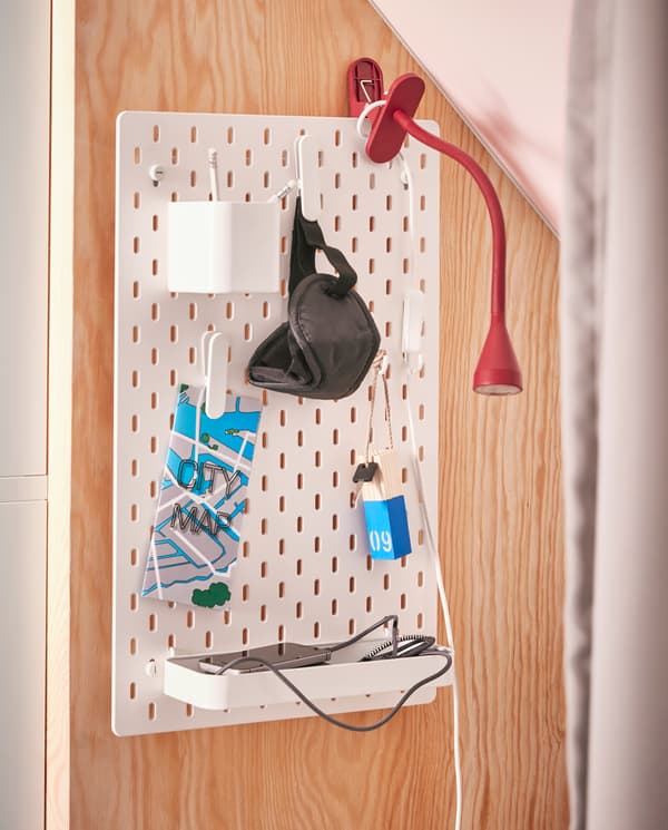 Zamocowana na ścianie biała tablica perforowana z zawieszonymi na haczykach drobnymi przedmiotami i czerwoną lampką z klipsem.