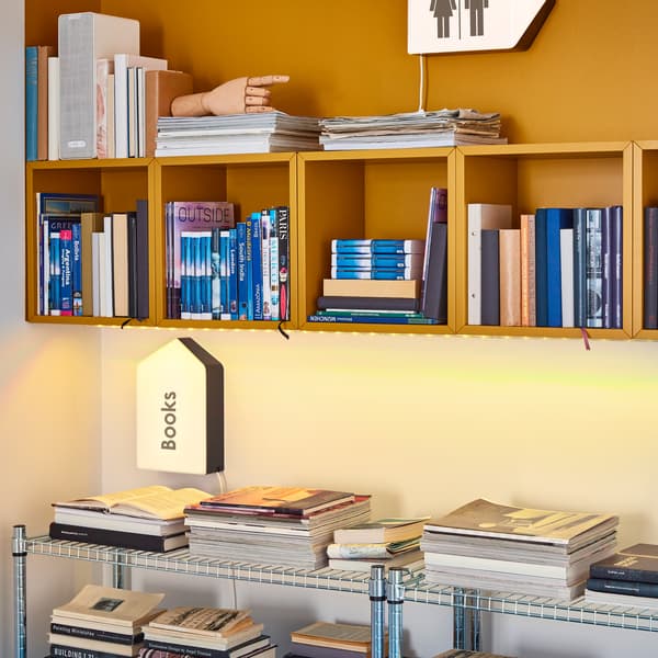 Zawieszone na ścianie, wypełnione książkami otwarte szafki EKET w kolorze żółtym i ustawione poniżej metalowe regały.