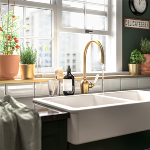 Zelena kuhinja s belim pločicama, kombinovana slavina boje mesinga, bela keramička sudopera i crnobraon saksija. 