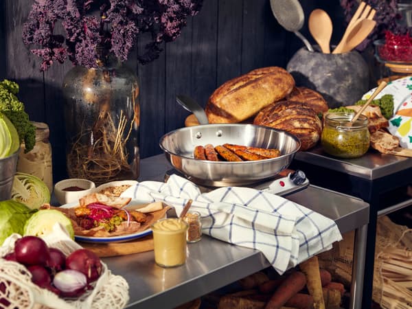 Zeleninové hot dogy KORVMOJ v pánvi, červená cibule, kapusta a další potraviny na nerezové pracovní ploše.