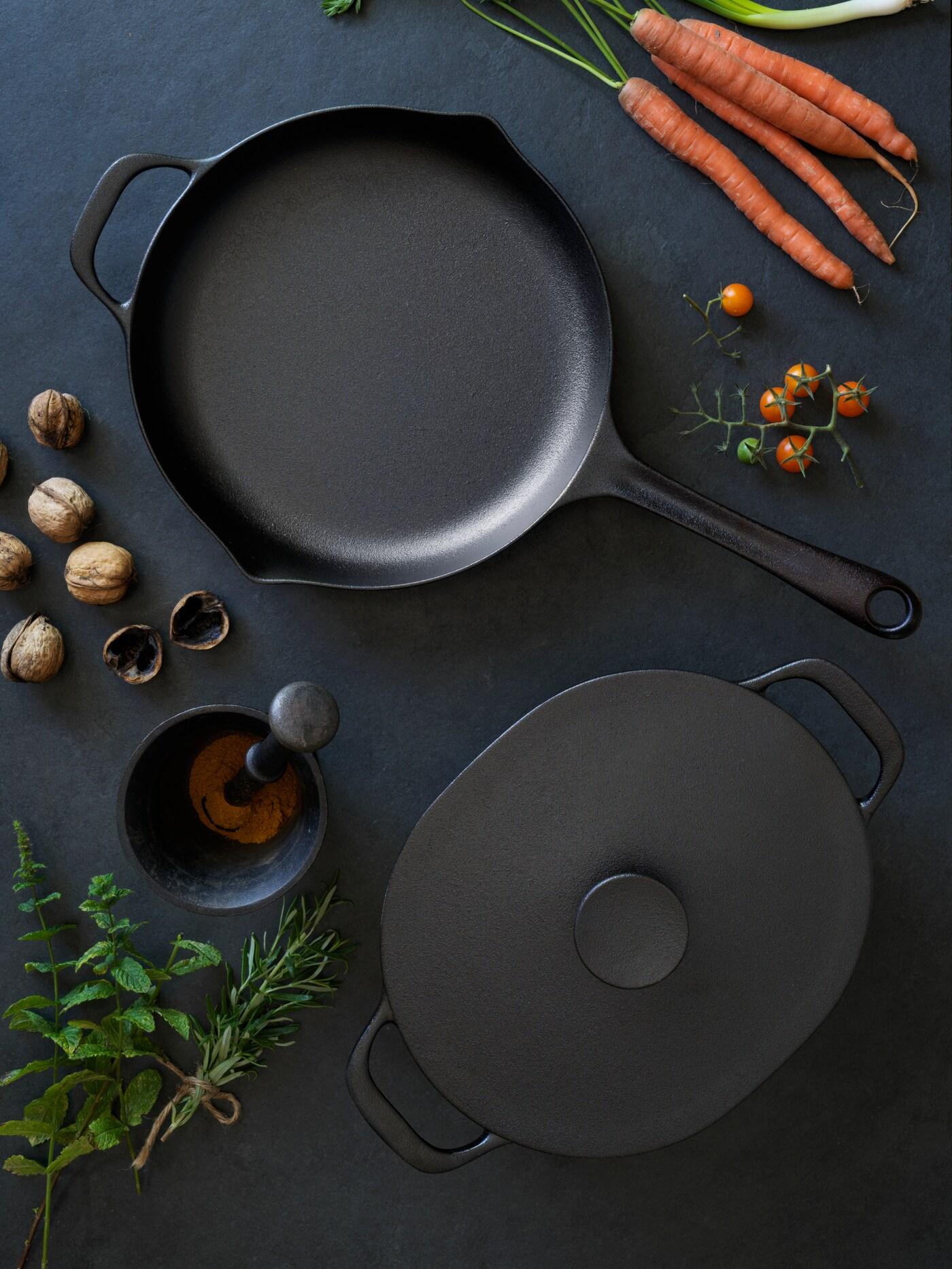 Żeliwna patelnia i garnek z serii VARDAGEN  leżą na czarnym blacie, dookoła leżą warzywa i przyprawy.