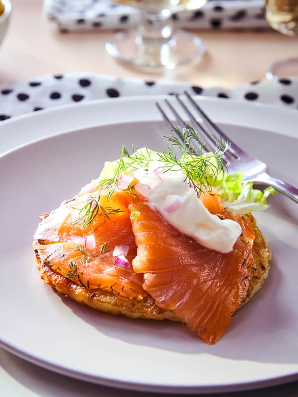 Zemiakové RÖSTI s údeným lososom SJÖRAPPORT, kyslou smotanou a kôprom na bielom tanieri, v pozadí sú biele servítky s čiernymi bodkami a dva poháre s bielym vínom.