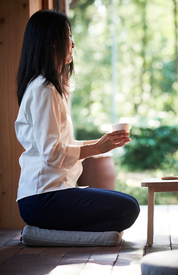 Žena drží v oboch rukách hrnček a sedí na podložke na sedenie položenej na zemi OMTÄNKSAM.