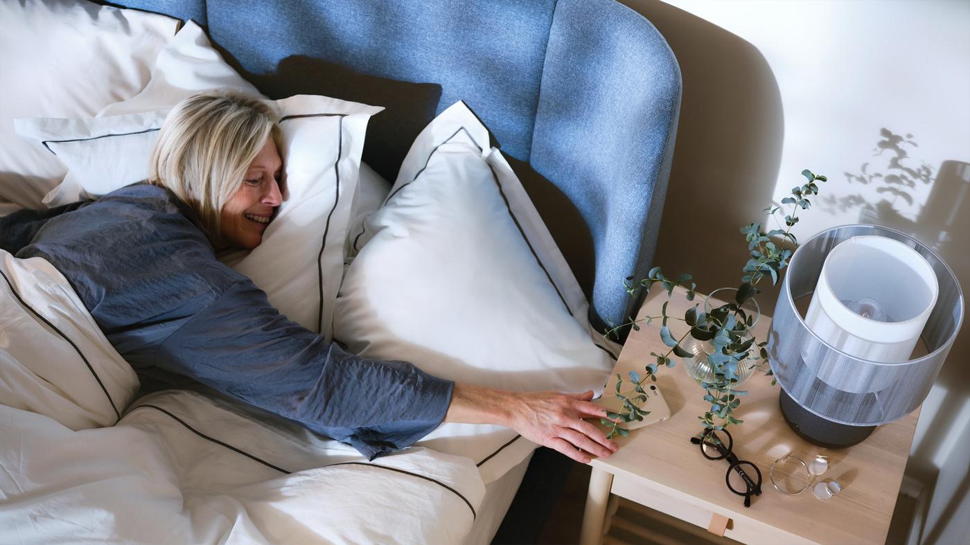 Žena leží v modrej čalúnenej posteli s bielymi obliečkami a naťahuje sa na drevený nočný stolík. Na stolíku je rastlina a reproduktorová lampa.