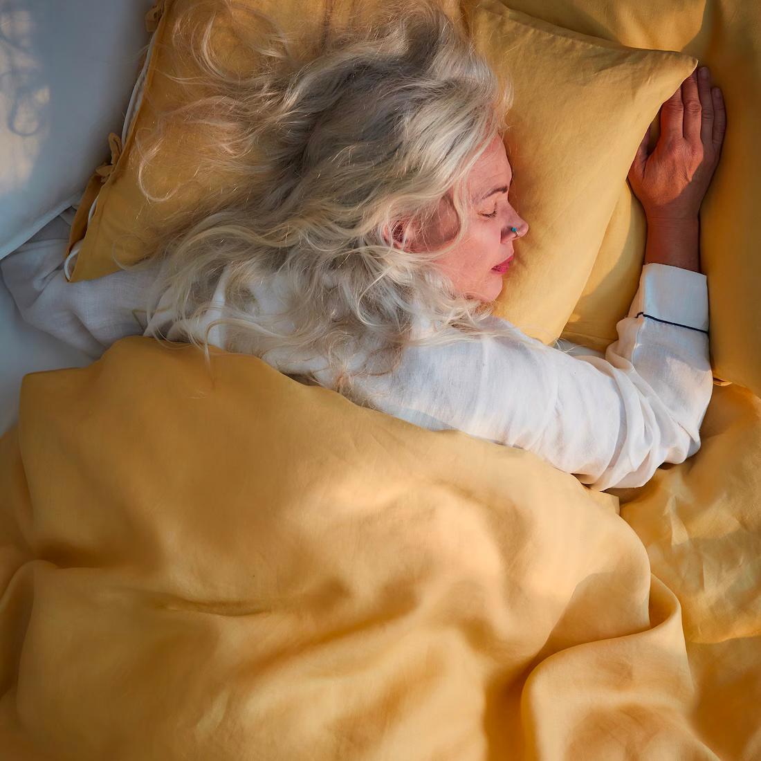 Žena ležící v pyžamu se světlými vlasy, přikrytá oranžově-žlutou přikrývkou a stejnobarevným polštářem pod její hlavou.