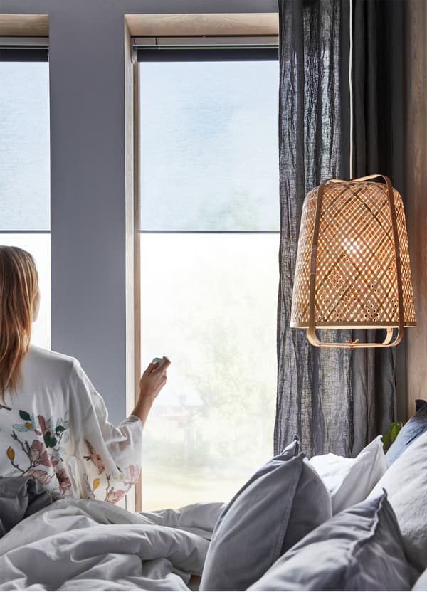 Žena sedi u spavaćoj sobi i daljinskim upravlja sivim IKEA KADRILJ rol-zastorima na baterije/bežičnim.