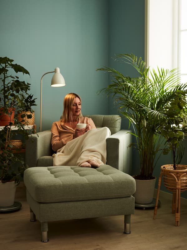 Žena sedí v kresle LANDKRONA so šálkou čaju v ruke obklopená rastlinami, niektoré z nich na stojane BUSKBO.