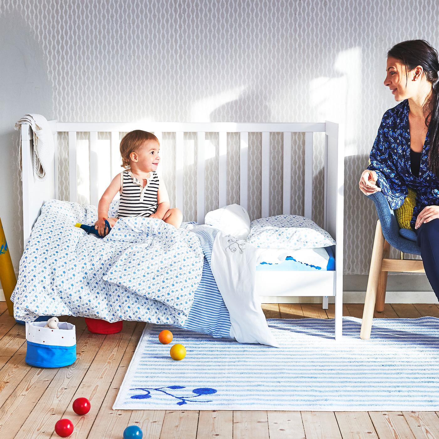 Žena sledujte bábätko na posteli s modro-bielymi posteľným textíliami GULSPARV. V popredí pásikavý koberec GULSPARV.
