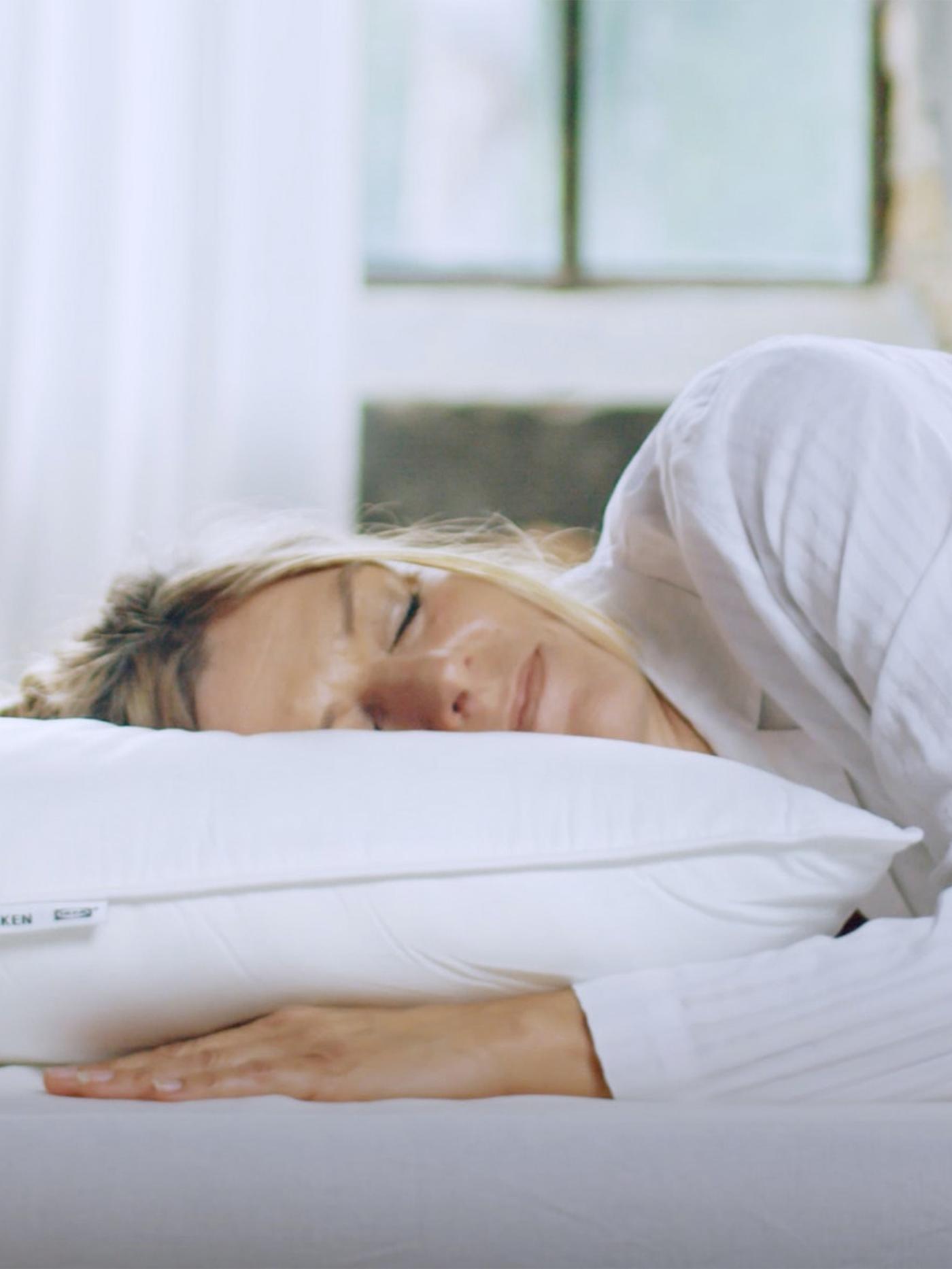 Žena spí na boku s hlavou na vysokém polštáři SKOGSFRÄKEN v bílém povlaku na polštář.